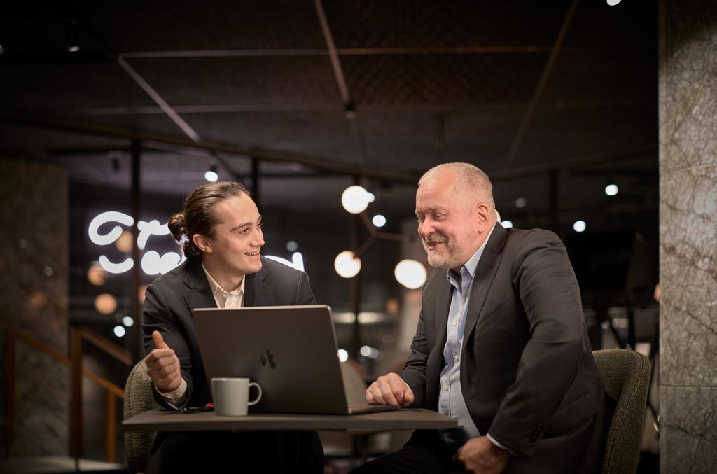 Två män i affärsklädsel sitter vid ett bord med en bärbar dator och en kaffekopp och för samtal i en modern, väl upplyst kontorsmiljö.